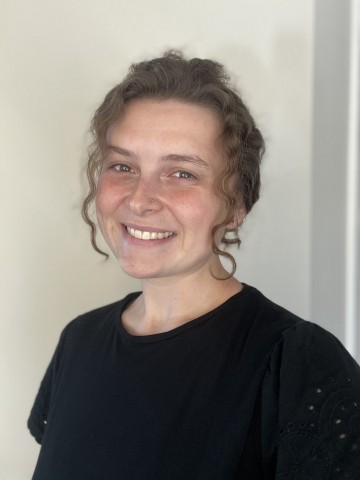 Photo of woman in a black shirt standing in front of a white background, smiling at the camera. 