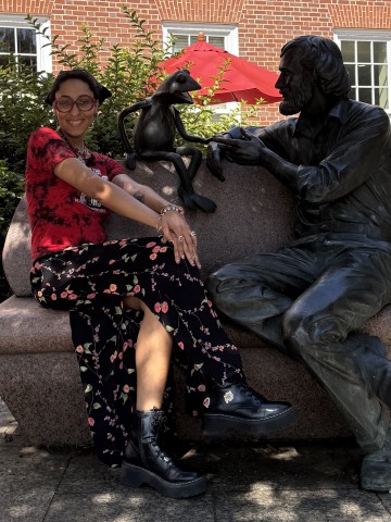 Young brown skinned person sitting on a bench smiling beside a statue of Kermit the Frog and Jim Henson. Person is wearing red circle glasses, a red and black tie-dye shirt and a long black skirt with floral patterning.