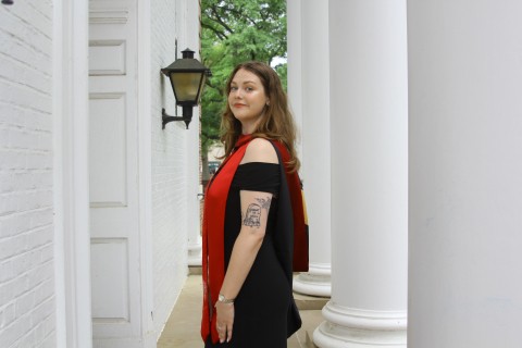 Woman with brown hair and tattoos in a black dress standing in front of Woods Hall on UMD Campus