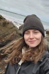 Picture shows a woman standing in front of a gray sea and black rocks. She has brown curly hair, is wearing a black beanie, and is smiling at the camera. 