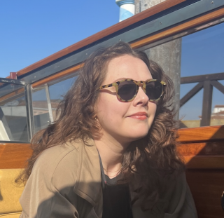 This is a photo of a woman with sunglasses and brown hair with a brown jacket on.